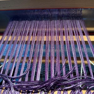 Puede incluir: Primer plano de un telar de madera con hilos morados y blancos. Los hilos est&aacute;n tejidos apretadamente, creando una superficie texturizada. El telar est&aacute; en proceso de crear un textil, con los hilos colgando en la parte inferior.