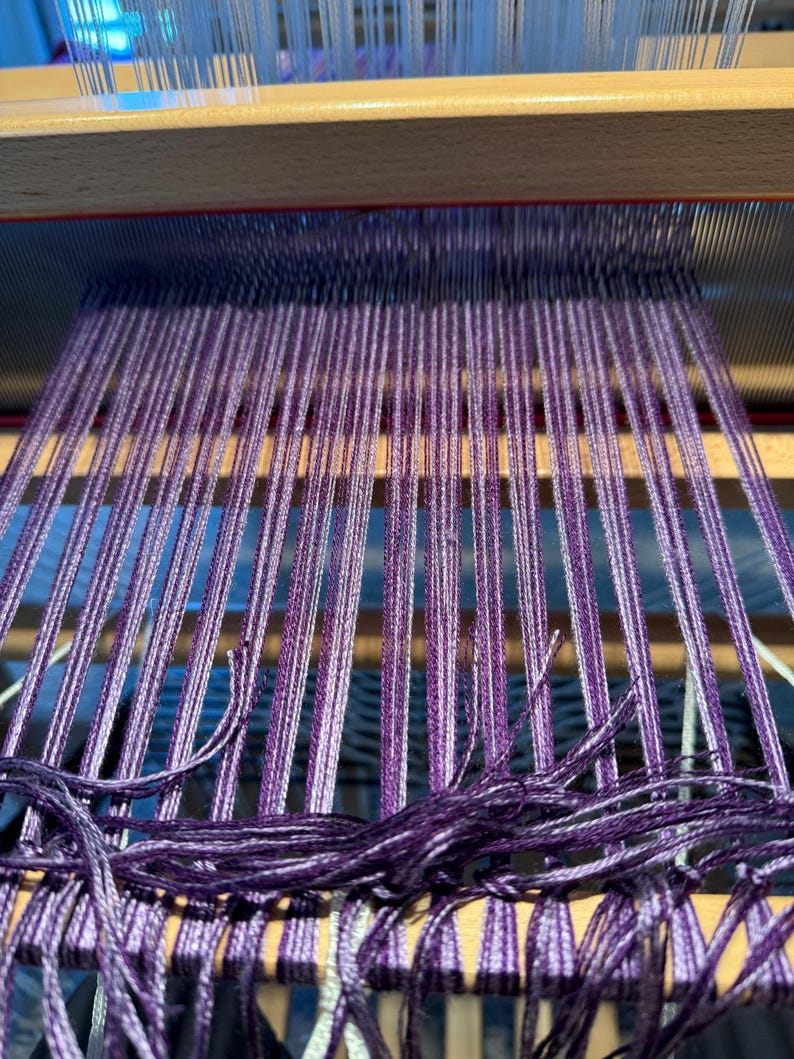 Puede incluir: Primer plano de un telar de madera con hilos morados y blancos. Los hilos est&aacute;n tejidos apretadamente, creando una superficie texturizada. El telar est&aacute; en proceso de crear un textil, con los hilos colgando en la parte inferior.