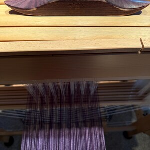 Puede incluir: Una lanzadera de tejido de madera en un tono p&uacute;rpura oscuro descansa sobre un telar de madera. La lanzadera est&aacute; llena de hilo p&uacute;rpura, lista para tejer. El telar presenta hilos verticales en tonos p&uacute;rpura y blanco.