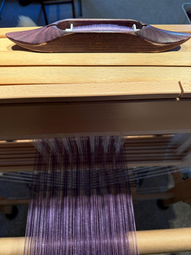 Puede incluir: Una lanzadera de tejido de madera en un tono p&uacute;rpura oscuro descansa sobre un telar de madera. La lanzadera est&aacute; llena de hilo p&uacute;rpura, lista para tejer. El telar presenta hilos verticales en tonos p&uacute;rpura y blanco.