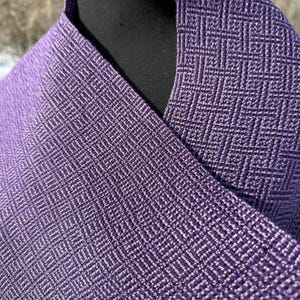 Puede incluir: Primer plano de una tela morada tejida con un patr&oacute;n de diamantes. La tela est&aacute; drapeada sobre una forma negra, mostrando la textura y las variaciones de color. El tejido crea una superficie texturizada.