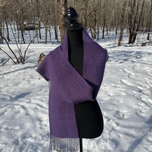 Puede incluir: Bufanda morada oscura con un dise&ntilde;o geom&eacute;trico y flecos. La bufanda est&aacute; colocada sobre un maniqu&iacute; negro, con un fondo nevado al aire libre. Se destacan la textura y el color de la bufanda.