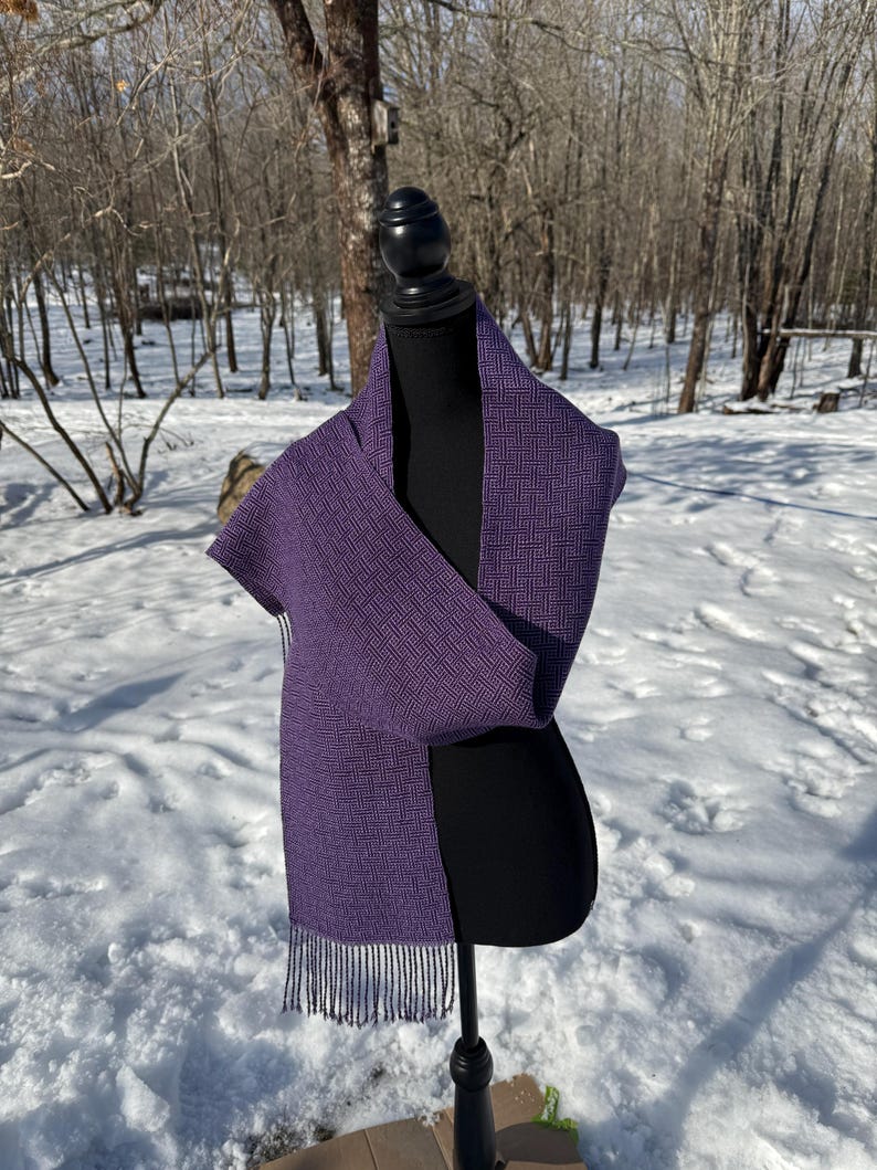 Puede incluir: Bufanda morada oscura con un dise&ntilde;o geom&eacute;trico y flecos. La bufanda est&aacute; colocada sobre un maniqu&iacute; negro, con un fondo nevado al aire libre. Se destacan la textura y el color de la bufanda.