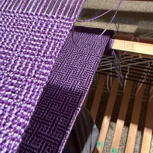 Puede incluir: Primer plano de un proyecto de tejido en curso, que muestra un patr&oacute;n geom&eacute;trico p&uacute;rpura y blanco. La tela tejida est&aacute; parcialmente terminada en un telar de madera, con hilos y la lanzadera visibles. La imagen destaca el proceso de tejido.