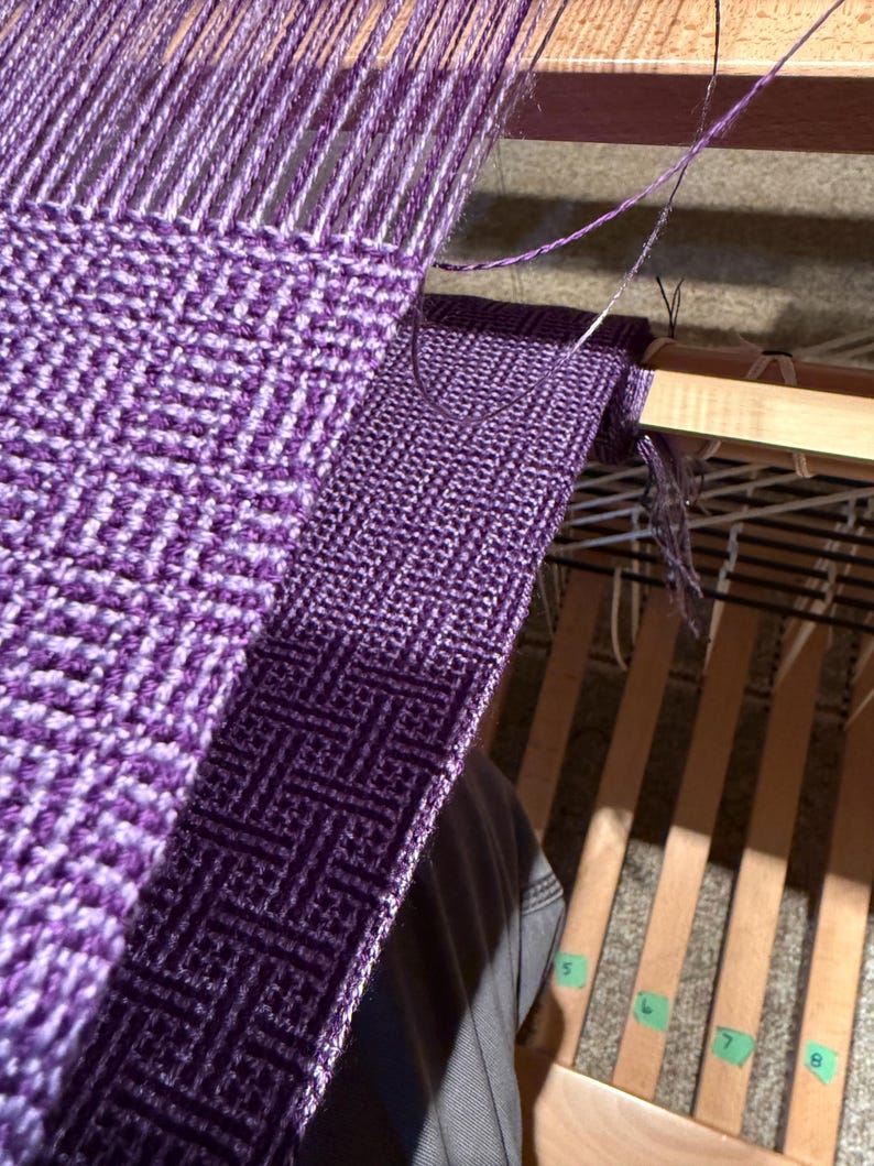 Puede incluir: Primer plano de un proyecto de tejido en curso, que muestra un patr&oacute;n geom&eacute;trico p&uacute;rpura y blanco. La tela tejida est&aacute; parcialmente terminada en un telar de madera, con hilos y la lanzadera visibles. La imagen destaca el proceso de tejido.