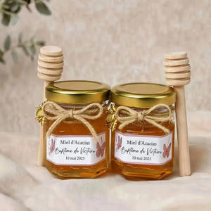 Peut inclure: Deux petits pots en verre remplis de miel doré, chacun avec un couvercle doré, un nœud en ficelle de jute et une breloque abeille. Les étiquettes indiquent Miel d'Acacias, Baptême de Victoire et 10 mai 2025. Chaque pot est accompagné d'une cuillère à miel en bois.