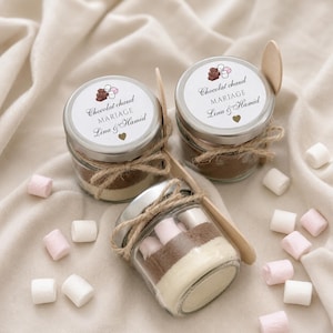 Op de afbeelding: Drie kleine glazen potjes gevuld met laagjes warme chocolademelkpoeder en marshmallows, elk met een zilveren deksel en een etiket met de tekst Chocolat chaud MARIAGE Lina & Hamid. Een houten lepel zit in het touw rond elk potje.