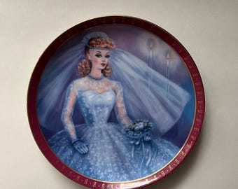 May include: A decorative plate featuring a bride in a blue wedding dress and veil, holding a bouquet. The plate has a maroon border with a gold pattern. The artwork is a painting of a woman with blonde hair and a blue necklace.