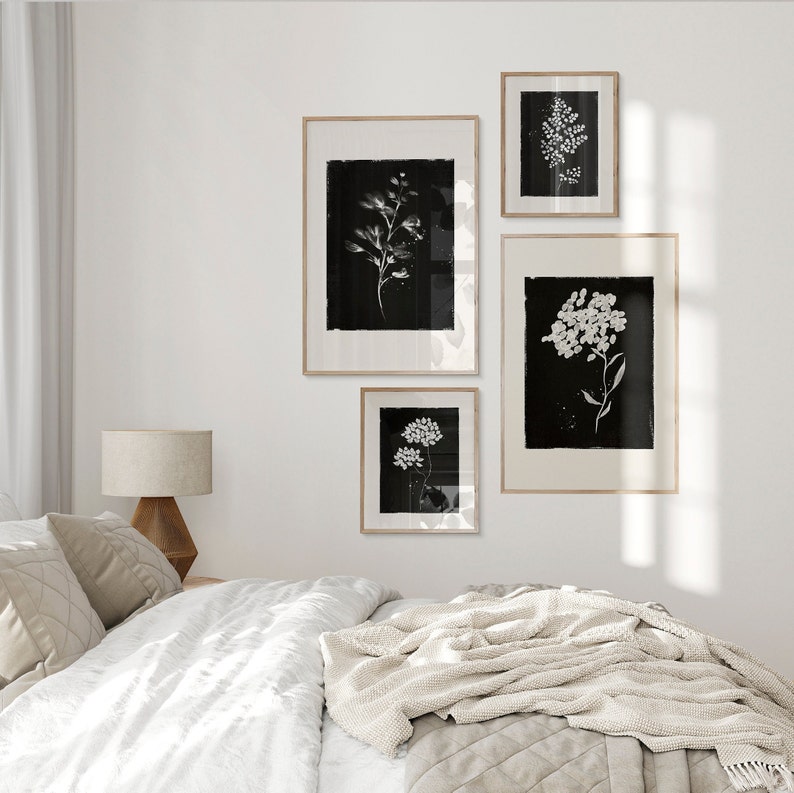 May include: Four black and white framed botanical prints with white flowers on a black background. The prints are hanging on a white wall above a white bed with a beige throw blanket.