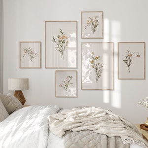 May include: A bedroom interior with a white wall and a bed with a white duvet and a beige throw blanket. There are six framed botanical prints on the wall featuring various flowers and plants in shades of pink, yellow, and green.
