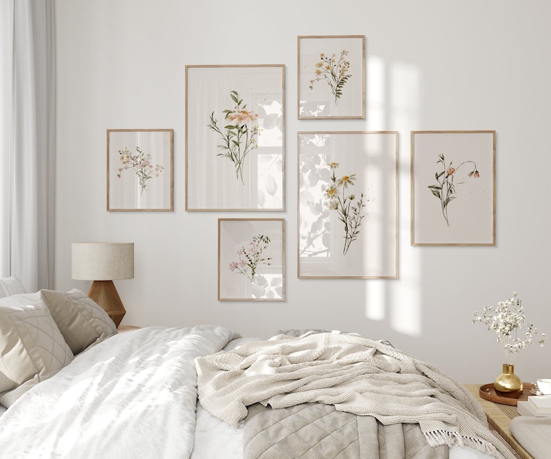 May include: A bedroom interior with a white wall and a bed with a white duvet and a beige throw blanket. There are six framed botanical prints on the wall featuring various flowers and plants in shades of pink, yellow, and green.