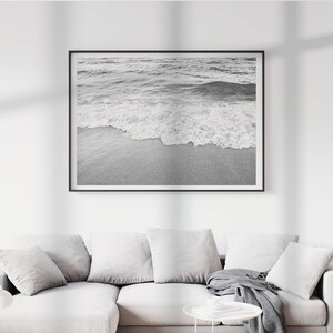 May include: A black and white photograph of a wave breaking on a sandy beach. The image is framed in a black frame and is hanging on a white wall.