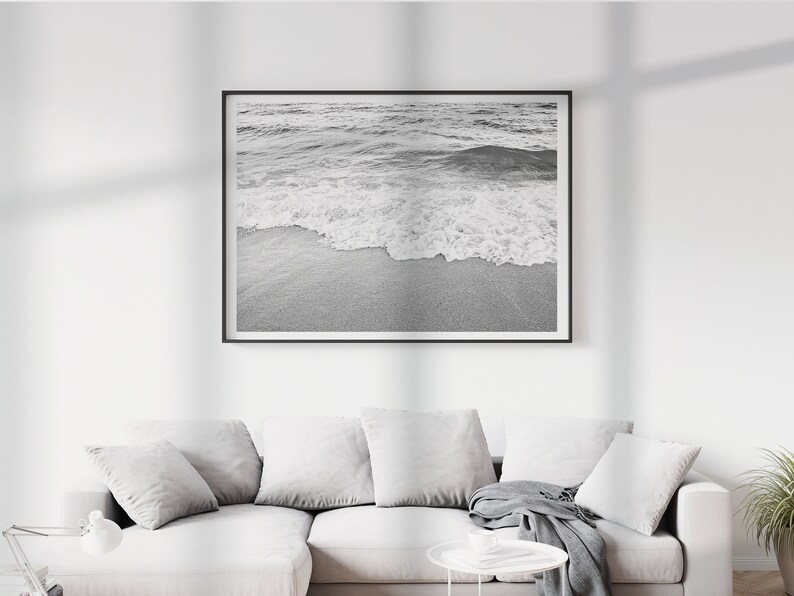 May include: A black and white photograph of a wave breaking on a sandy beach. The image is framed in a black frame and is hanging on a white wall.