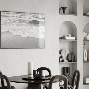 May include: A black and white photograph of ocean waves crashing on a sandy beach hangs on a white wall above a black marble dining table with four chairs. The table is decorated with a white vase, a black vase, and a black candle holder.