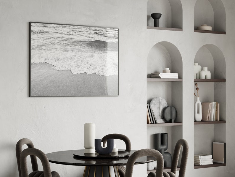 May include: A black and white photograph of ocean waves crashing on a sandy beach hangs on a white wall above a black marble dining table with four chairs. The table is decorated with a white vase, a black vase, and a black candle holder.