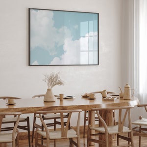 May include: A wooden dining table with six chairs, a vase with dried flowers, and a framed print of a blue sky with clouds. The table is made of light wood and has a natural finish. The chairs are also made of wood and have a light brown finish. The print is in a black frame and is hanging on a white wall.