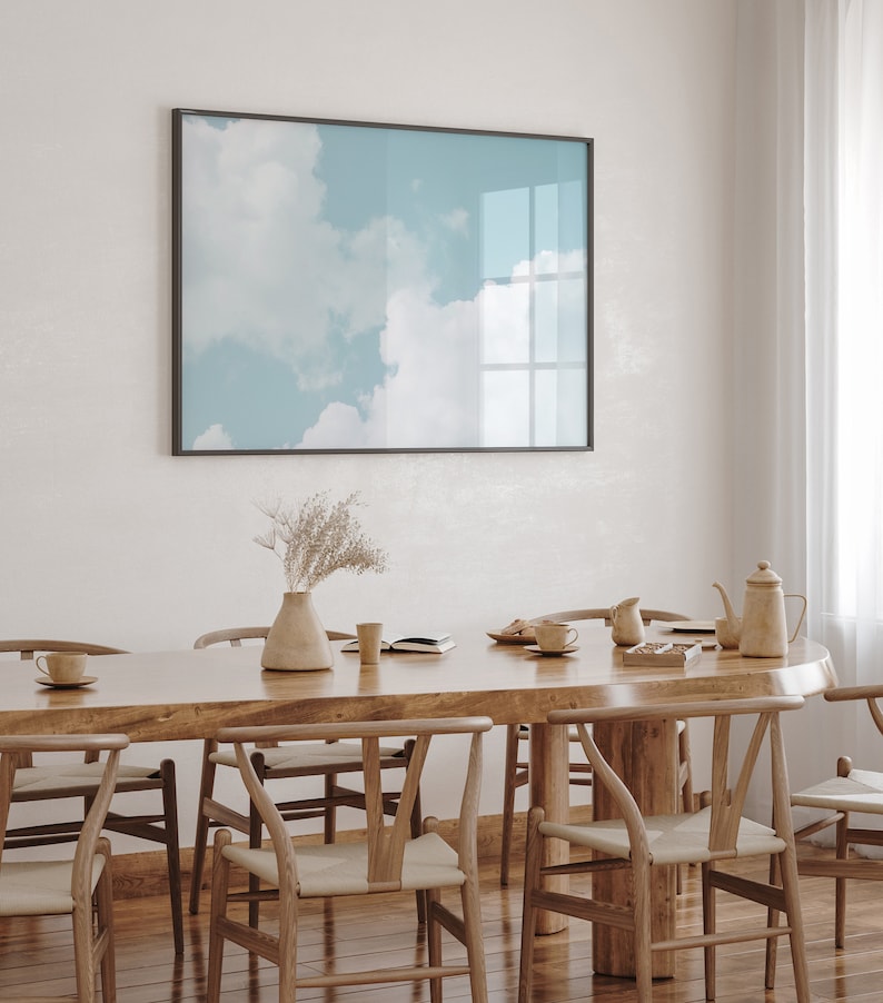 May include: A wooden dining table with six chairs, a vase with dried flowers, and a framed print of a blue sky with clouds. The table is made of light wood and has a natural finish. The chairs are also made of wood and have a light brown finish. The print is in a black frame and is hanging on a white wall.