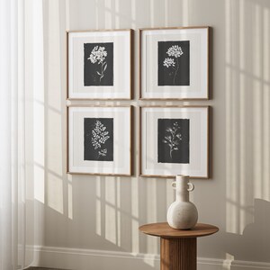 May include: Four framed black and white botanical prints with white flowers and leaves on a black background. The prints are arranged in a grid pattern on a light beige wall. A white ceramic vase with two handles sits on a wooden table in front of the prints.
