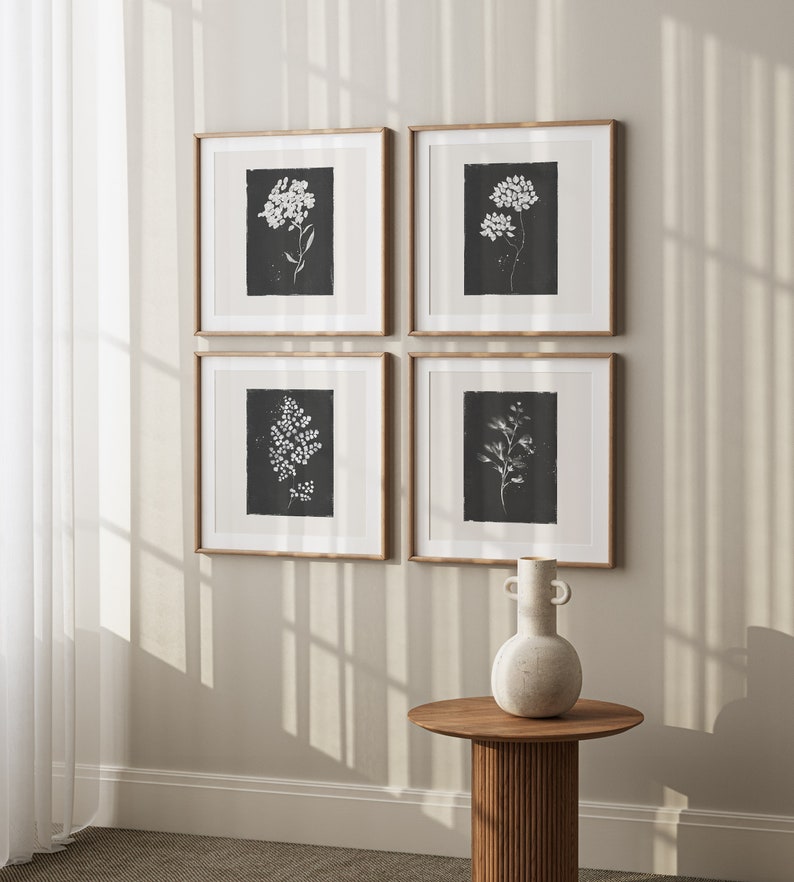May include: Four framed black and white botanical prints with white flowers and leaves on a black background. The prints are arranged in a grid pattern on a light beige wall. A white ceramic vase with two handles sits on a wooden table in front of the prints.