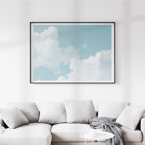 May include: A framed photograph of a blue sky with white clouds. The photograph is hanging on a white wall above a white couch with gray pillows and a gray throw blanket.