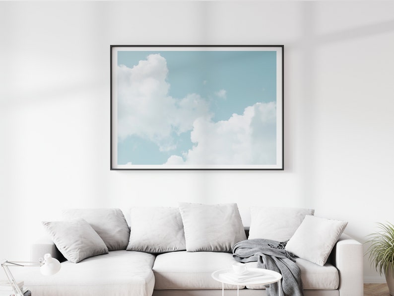 May include: A framed photograph of a blue sky with white clouds. The photograph is hanging on a white wall above a white couch with gray pillows and a gray throw blanket.