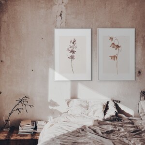 May include: Two framed watercolor prints of botanical illustrations. The print on the left features a single stem of purple flowers. The print on the right features a single stem of brown flowers. The prints are hanging on a light brown wall above a bed with white bedding.