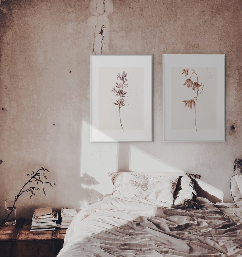 May include: Two framed watercolor prints of botanical illustrations. The print on the left features a single stem of purple flowers. The print on the right features a single stem of brown flowers. The prints are hanging on a light brown wall above a bed with white bedding.