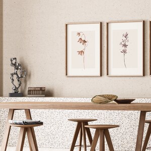May include: Two framed botanical prints with delicate brown and purple flowers on a light beige background, hanging above a wooden table with three wooden stools. A small metal sculpture sits on a shelf below the prints.