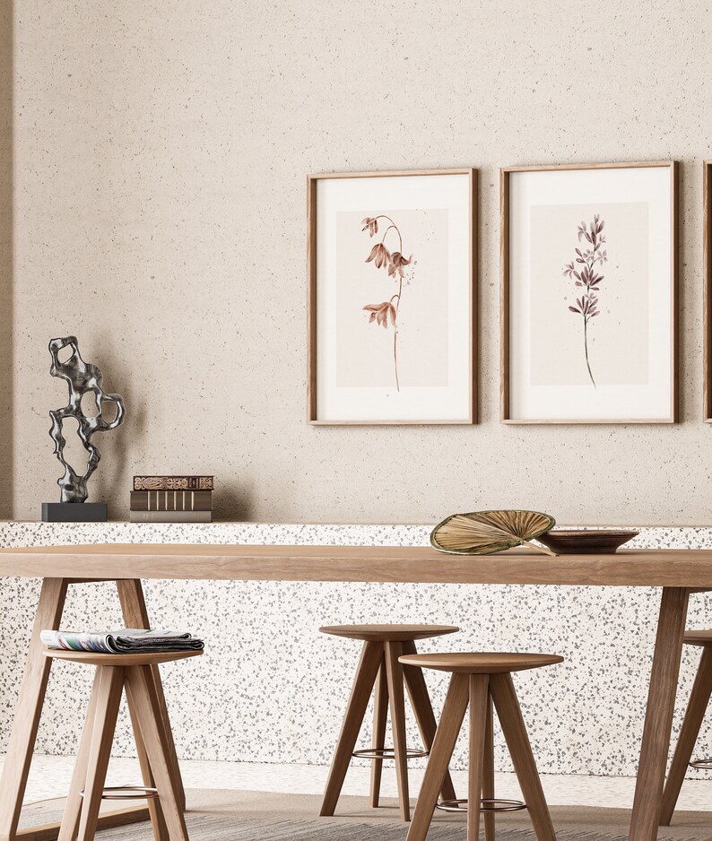 May include: Two framed botanical prints with delicate brown and purple flowers on a light beige background, hanging above a wooden table with three wooden stools. A small metal sculpture sits on a shelf below the prints.