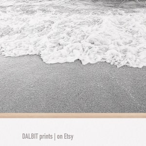 May include: Black and white photograph of a sandy beach with foamy waves crashing on the shore.