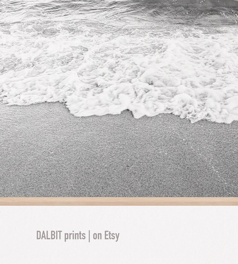 May include: Black and white photograph of a sandy beach with foamy waves crashing on the shore.
