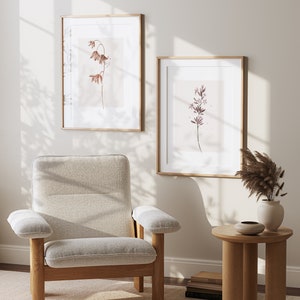 May include: Two framed botanical prints with delicate brown and purple flowers on a light background. The prints are hanging on a white wall with a beige armchair and a small wooden side table in the foreground.