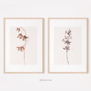 May include: Two framed watercolor prints of botanical illustrations. The print on the left features a brown flower with a delicate stem. The print on the right features a purple flower with a delicate stem.