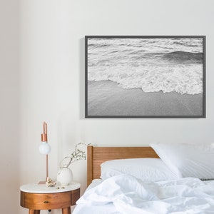 May include: Black and white photograph of a wave breaking on a sandy beach. The image is framed in a black frame and hangs on a white wall above a bed with white bedding.