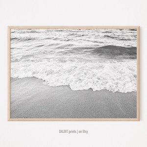 May include: Black and white photograph of a wave breaking on a sandy beach. The image is framed in a light wood frame.