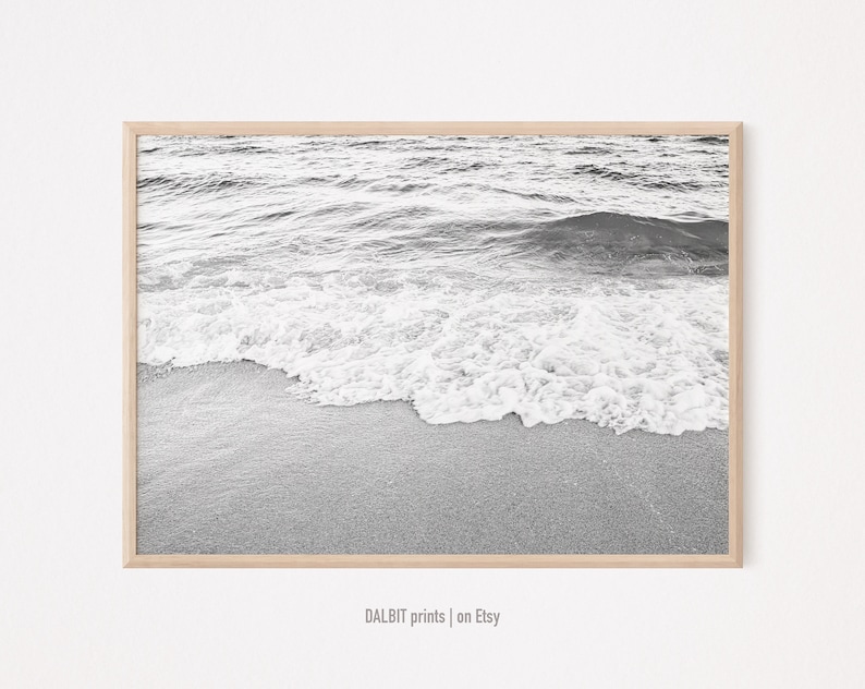 May include: Black and white photograph of a wave breaking on a sandy beach. The image is framed in a light wood frame.