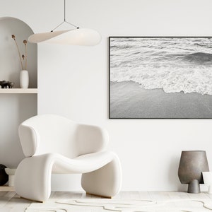 May include: A white armchair with a curved back and a white, wavy rug in front of it. A black and white photograph of ocean waves crashing on a sandy beach hangs on the wall. A white shelf with books and decorative objects is on the left side of the image.