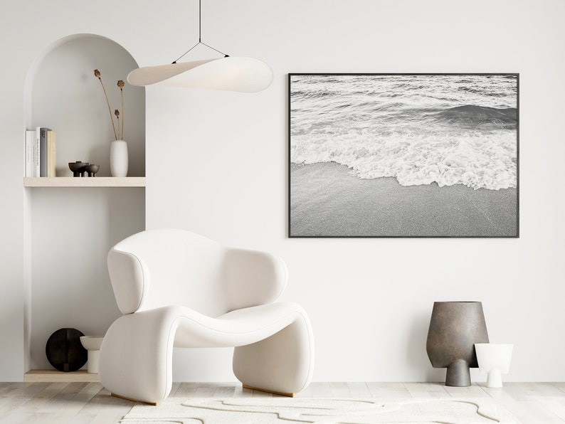May include: A white armchair with a curved back and a white, wavy rug in front of it. A black and white photograph of ocean waves crashing on a sandy beach hangs on the wall. A white shelf with books and decorative objects is on the left side of the image.