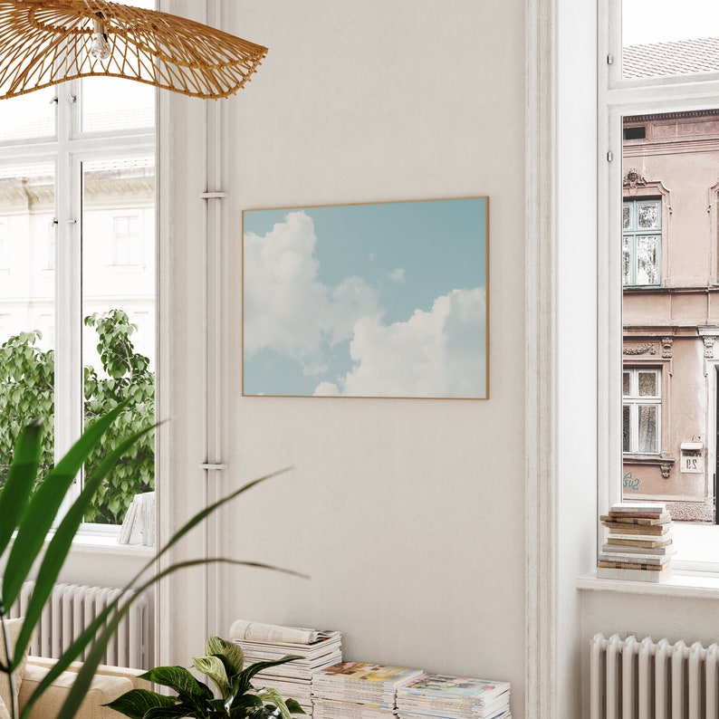 May include: A framed print of a blue sky with white clouds. The print is hanging on a white wall in a room with a window and a wooden ceiling fan.