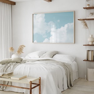 May include: A white bed with a light gray throw blanket and a wooden bench in front of it. A framed print of a blue sky with white clouds hangs on the wall behind the bed.