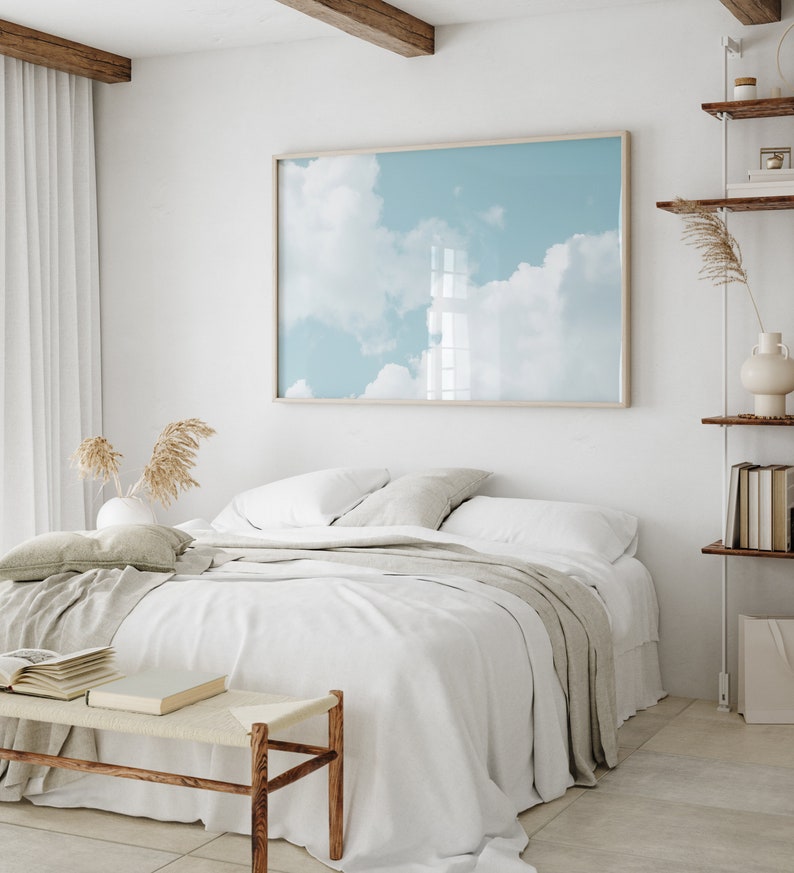 May include: A white bed with a light gray throw blanket and a wooden bench in front of it. A framed print of a blue sky with white clouds hangs on the wall behind the bed.