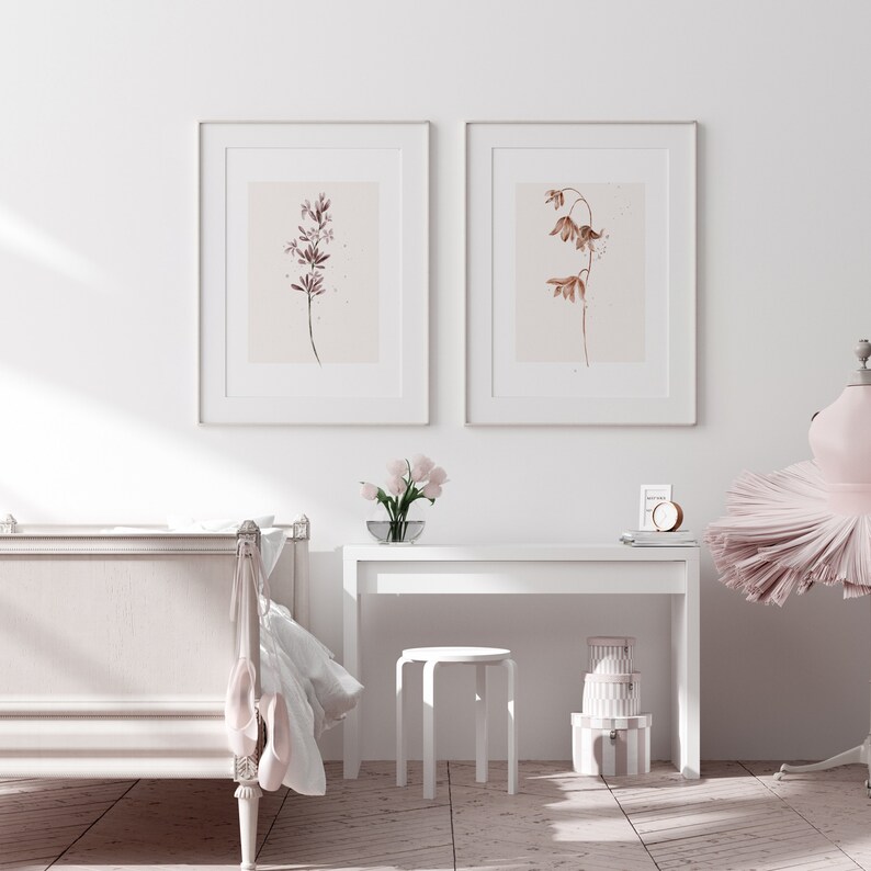 May include: Two framed watercolor prints of botanical illustrations. The print on the left features a pink flower with a brown stem. The print on the right features a brown flower with a brown stem. The prints are hanging on a white wall above a white desk with a white stool.