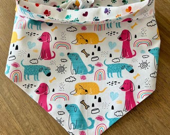 May include: A white dog bandana with a colorful pattern of dogs, rainbows, and clouds. The bandana is tied in a knot at the top.