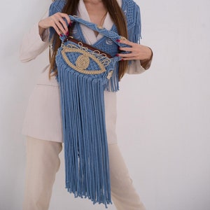 May include: A blue crocheted bag with a large eye design and fringe. The bag is held by a person wearing a beige blazer and pants.
