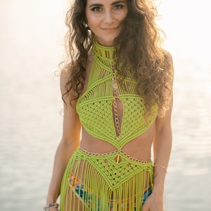 May include: A woman wearing a green crocheted halter top and skirt with fringe. The top features a gold chain detail and the skirt has a gold chain belt. The woman is standing in front of a body of water.