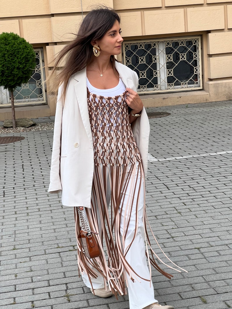 May include: A white blazer with a brown and white crocheted fringe top. The top has a white tank top underneath. The person is wearing white trousers and brown shoes.