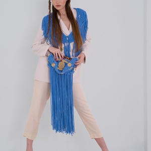 May include: A woman wearing a white blazer with a blue crocheted fringe shawl and matching bag. The bag has a woven design with brown accents.