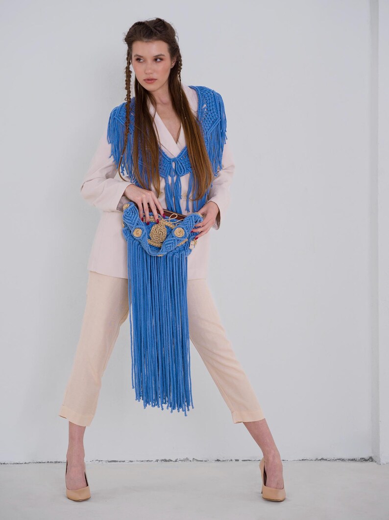 May include: A woman wearing a white blazer with a blue crocheted fringe shawl and matching bag. The bag has a woven design with brown accents.