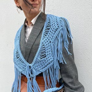 May include: A woman wearing a gray blazer with a light blue crocheted fringe shawl and a matching light blue crocheted bag with gold accents.