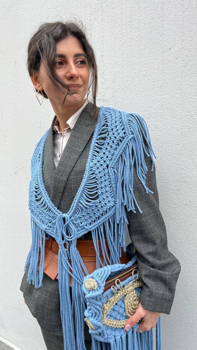 May include: A woman wearing a gray blazer with a light blue crocheted fringe shawl and a matching light blue crocheted bag with gold accents.